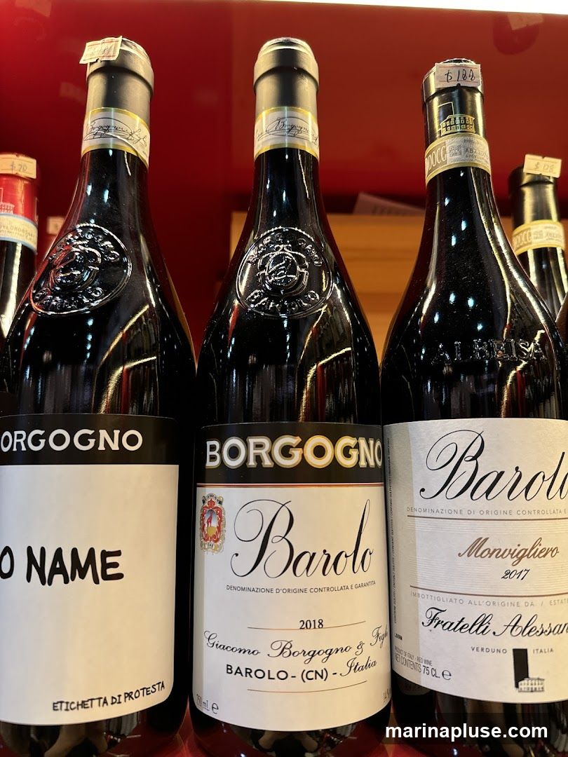 Three bottles of red wine are displayed side-by-side. The bottles have dark glass and prominent labels. The leftmost bottle has a label with "BORGOGNO" at the top and "O NAME" in large letters below. The middle bottle also has "BORGOGNO" at the top and "Barolo" in script below, with details about the year and producer. The rightmost bottle has "Barolo" in script and information about "Monvigliero 2017" and "Fratelli Alessandri". Price tags are visible on the necks of the bottles.
