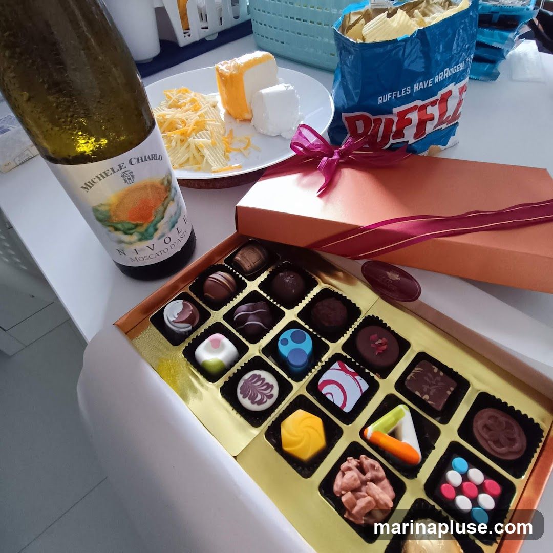 A box of assorted chocolates, a bottle of Michele Chiarlo Nivole Moscato d'Asti wine, a plate with cheese and shredded cheese, and a bag of Ruffles potato chips are arranged on a white surface.