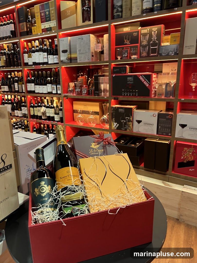 A red gift basket filled with straw contains two bottles of wine, one of which is Veuve Clicquot champagne. There is also a jar of what appears to be jam or preserves. A yellow box with an illustration of wine glasses is also in the basket. The background shows shelves stocked with various bottles of wine and spirits, as well as gift boxes and glassware.