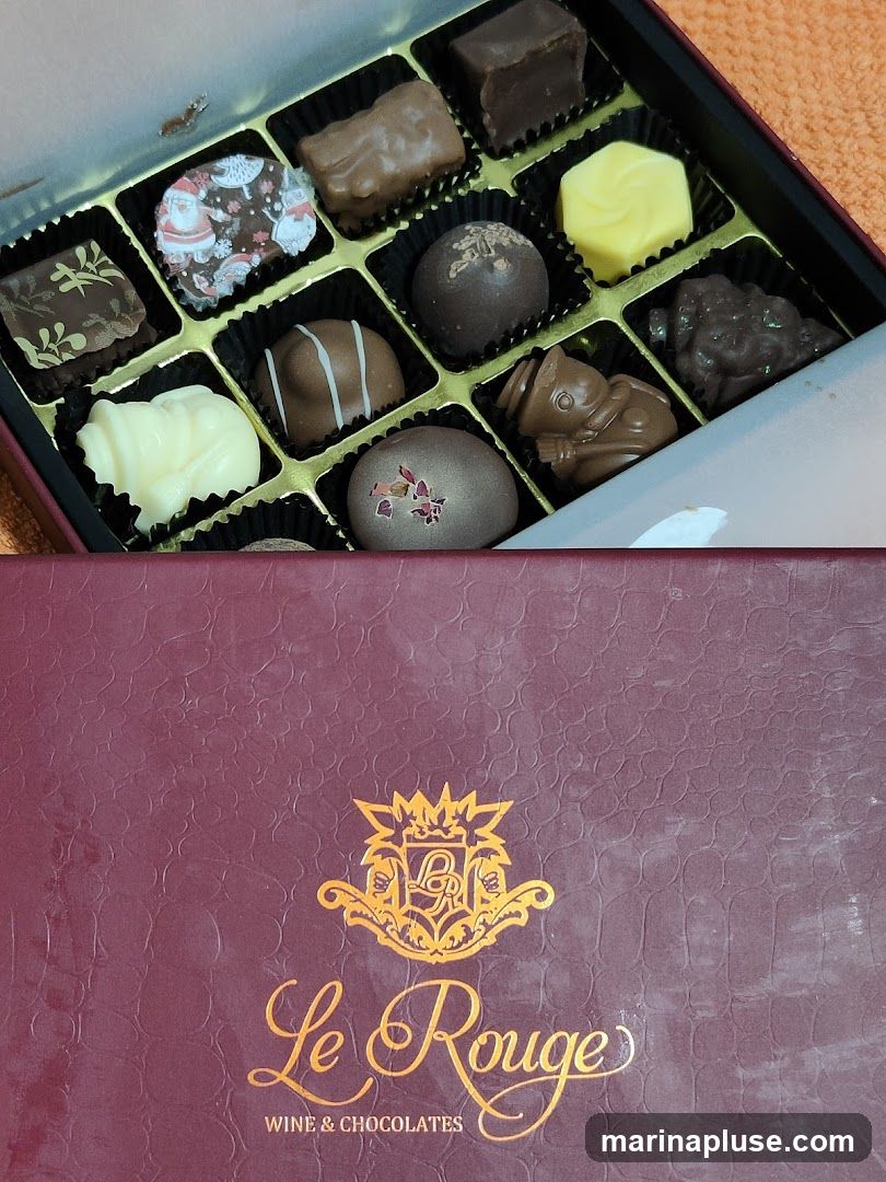 A box of assorted chocolates with various shapes and designs. Some chocolates have festive patterns, while others are plain or molded into figures. The box is partially open, revealing the chocolates arranged in individual compartments. The lid of the box, which is a deep burgundy color, is visible at the bottom of the image and features a gold embossed logo and the text "Le Rouge WINE & CHOCOLATES" in elegant script.