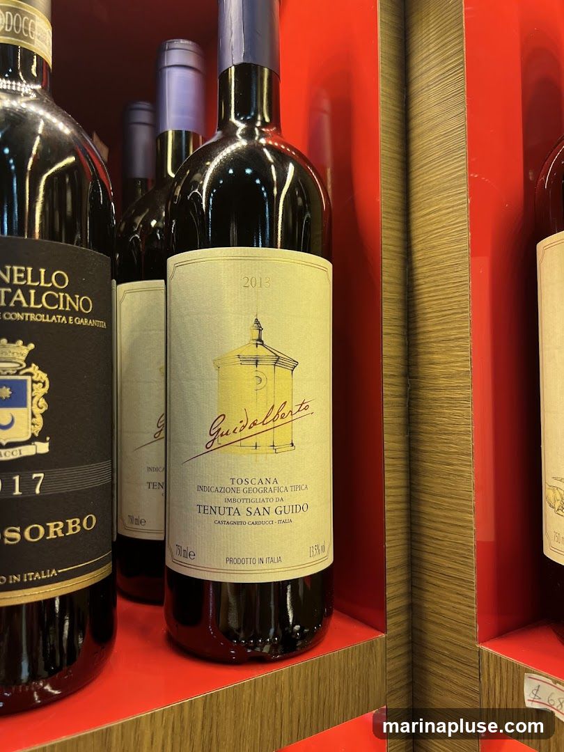 Several bottles of red wine are displayed on red shelves. The bottle in the foreground has a label that reads "Guidalberto Toscana Indicazione Geografica Tipica Imbottigliato da Tenuta San Guido Castagneto Carducci - Italia 2013". Another bottle to the left has a label that reads "Brunello di Montalcino Controllata e Garantita 2017". A price tag of $68 is visible at the bottom right.