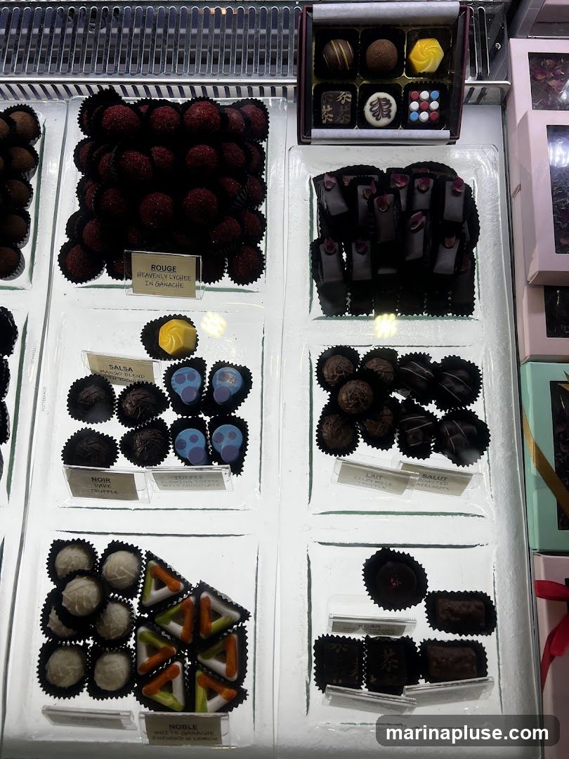 A display case filled with various types of chocolates. There are rows of dark chocolate truffles, red-coated lychee chocolates, and white chocolates. Some chocolates are decorated with colorful patterns or edible flowers. Small labels indicate some of the flavors, such as "Rouge Heavenly Lychee in Ganache," "Noir Dark Truffle," and "Noble White Ganache Infused w Lemon."