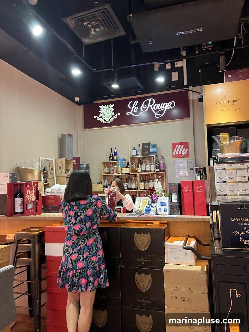 A woman in a floral dress stands at a counter in a store selling wine and spirits. Behind the counter, shelves are stocked with various bottles of alcohol. A sign above reads "Le Rouge". Boxes of Dom Perignon and Veuve Clicquot are stacked near the counter.