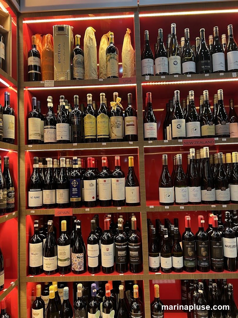 A wall of shelves filled with various bottles of wine. The shelves are red and illuminated by strip lights. Different sections are labeled with country names like 'USA', 'SPAIN', and 'NEW ZEALAND'. Bottles are arranged in rows, with some bottles in gift boxes or bags.
