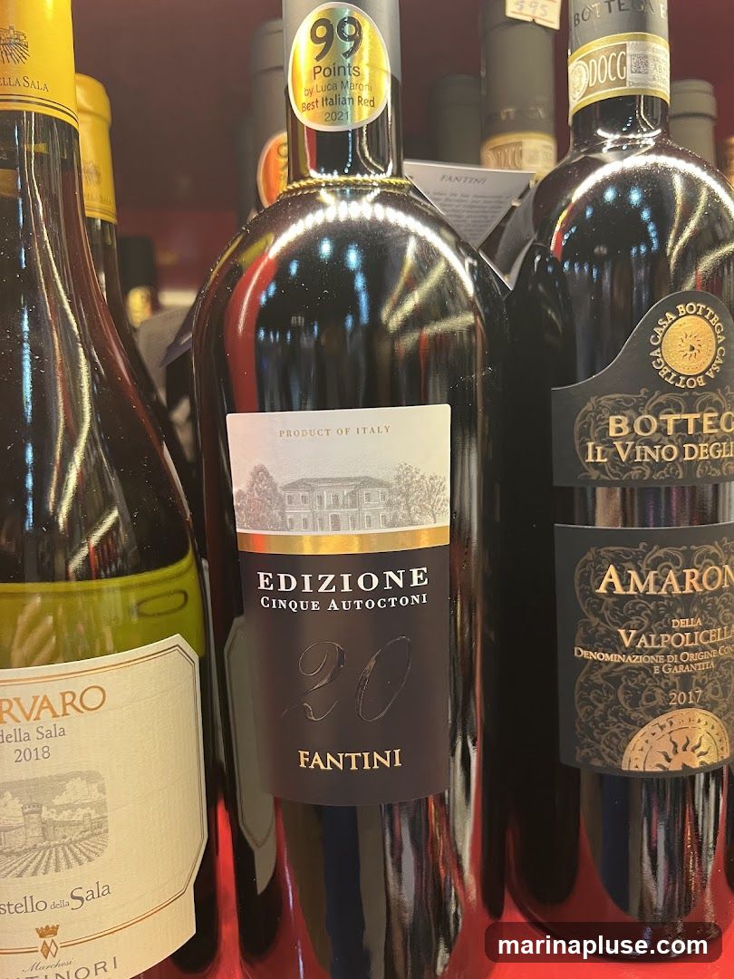 Several bottles of Italian red wine are displayed on a shelf. One bottle in the center is labeled "EDIZIONE CINQUE AUTOCTONI 20 FANTINI" and has a sticker indicating "99 Points by Luca Maroni Best Italian Red 2021". To its right is a bottle of "AMARONE DELLA VALPOLICELLA 2017". To the left is a bottle labeled "RVARO della Sala 2018" with a yellow cap.
