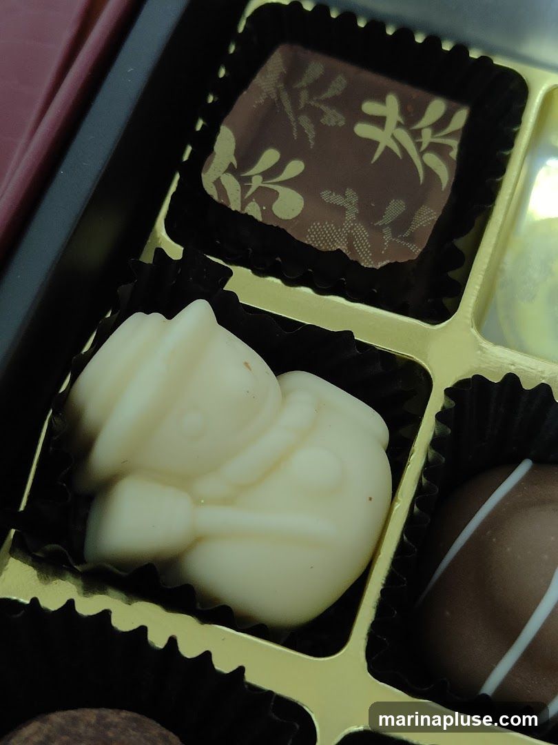 A close-up view of a box of assorted chocolates. In the foreground, a white chocolate truffle shaped like a snowman with a broom is visible. To its right, a dark chocolate truffle with white drizzle is partially seen. Above the snowman, a dark chocolate square with a gold leaf pattern is displayed. The chocolates are arranged in a gold-lined box with black paper dividers.