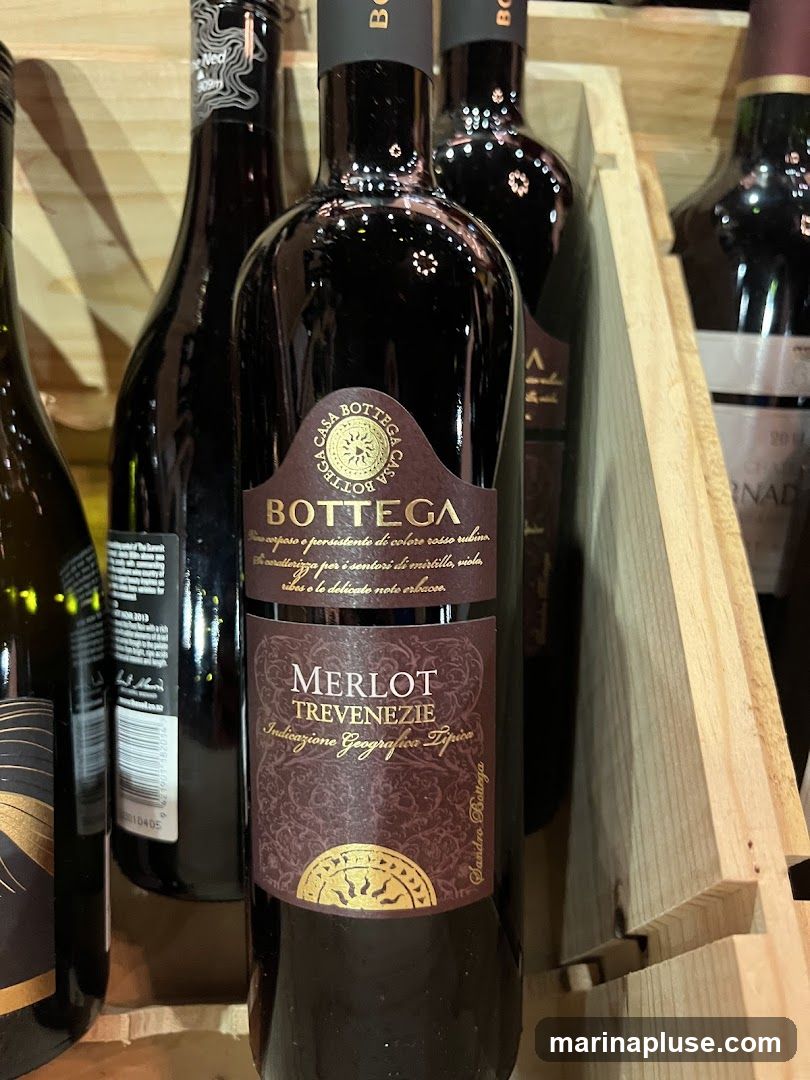 Several bottles of Bottega Merlot Trevenezie wine are displayed in a wooden crate. The central bottle is in focus, showing its label with the brand name "BOTTEGA" and details about the wine, including "MERLOT TREVENEZIE" and "Indicazione Geografica Tipica". Other bottles are partially visible in the background.