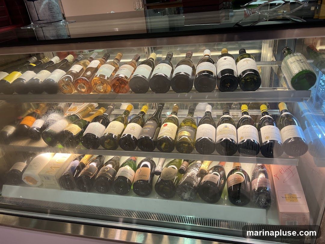 A refrigerated display case filled with various bottles of wine and champagne. The bottles are arranged on three shelves, with some bottles standing upright and others lying on their sides. The display case is well-lit, and the labels on the bottles are visible.