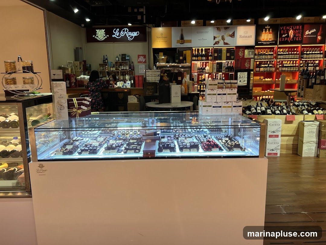 A well-lit interior of a shop selling chocolates and wine. In the foreground, a glass display case showcases an assortment of chocolates. Behind it, shelves are stocked with various bottles of wine and other beverages. A counter area with staff is visible in the background, along with branded signage for "Le Rouge" and other wine labels.