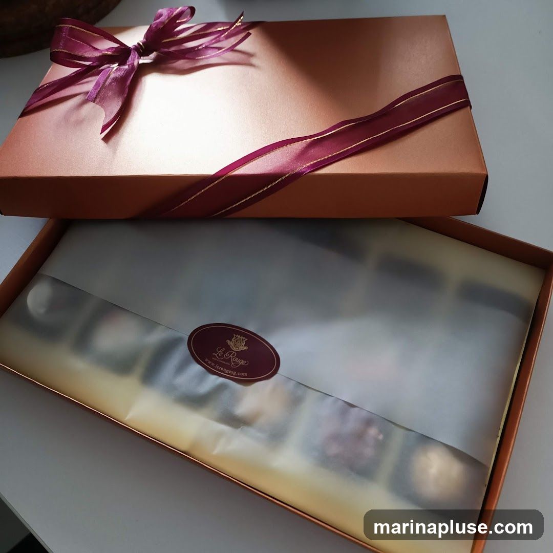 An open box of chocolates, tied with a burgundy ribbon. The box is copper-colored and contains an assortment of chocolates covered in white tissue paper. A sticker with the logo "Le Rouge" and website "www.lerougesg.com" is visible on the tissue paper.