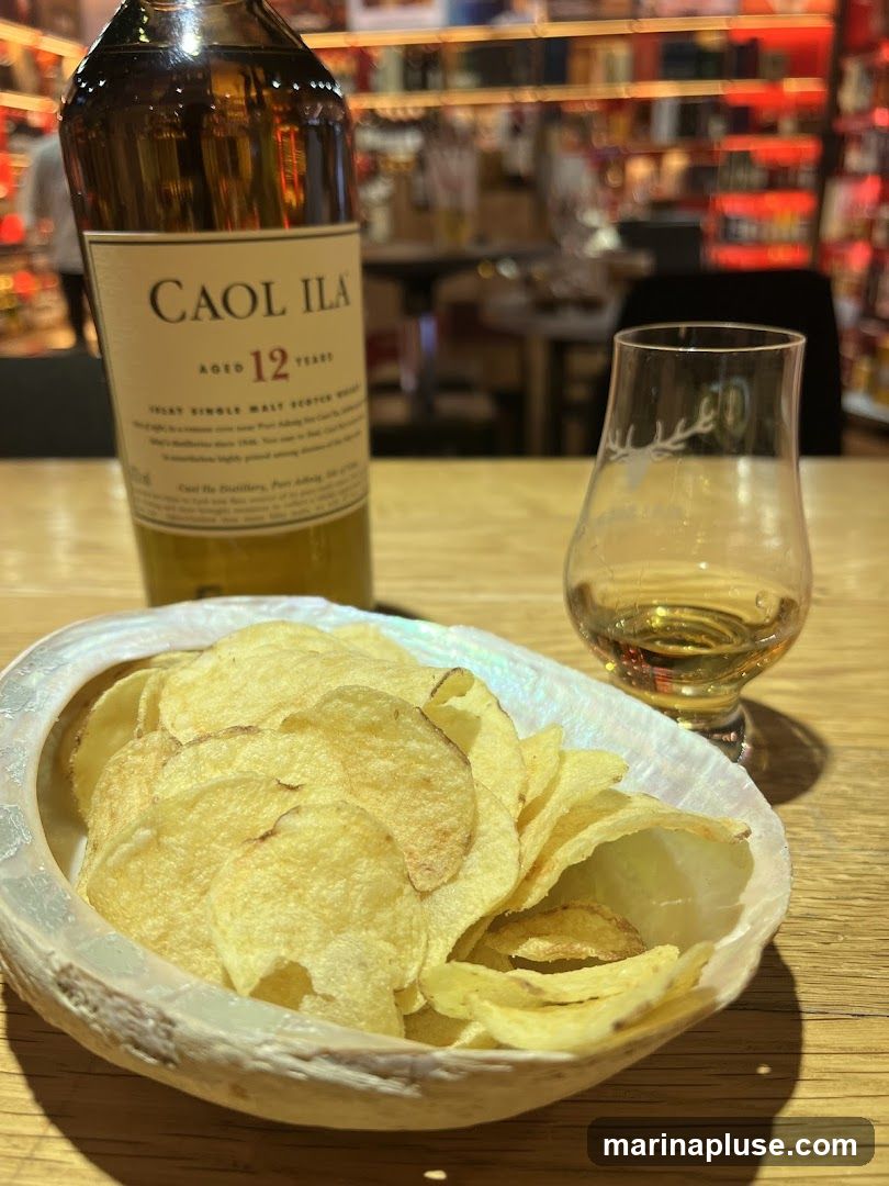 A bottle of Caol Ila Aged 12 Years Scotch Whisky sits next to a tasting glass with a small amount of amber liquid. In front of the glass is a bowl of potato chips served in a large shell. The background shows a blurred view of a bar or shop with shelves stocked with bottles.
