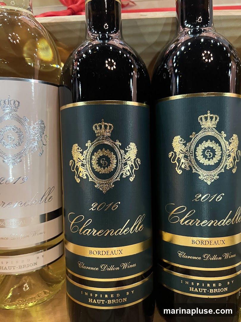 Close-up of several bottles of Clarendelle wine. The bottles on the right are dark red wine, and the bottle on the left appears to be white wine. The labels are dark green with gold lettering and feature a crest with lions and a crown. The labels indicate the year, wine name, region (Bordeaux), producer (Clarence Dillon Wines), and that it is inspired by Haut-Brion.