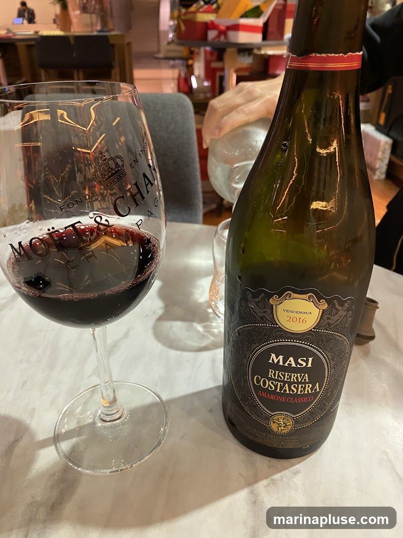 A bottle of Masi Riserva Costasera Amarone Classico wine from 2016 sits next to a glass of red wine. The glass has the Moët & Chandon logo visible. The setting appears to be indoors, possibly a restaurant or home, with blurred background elements including gift boxes.