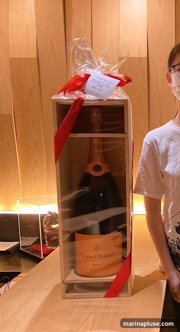 A large bottle of Veuve Clicquot Brut champagne is presented in a wooden box, wrapped in clear plastic and tied with red ribbon. A small card attached to the ribbon reads "ALL THE BEST Happy Anniversary". A person wearing glasses and a face mask stands to the right of the gift. The background features a wooden slatted wall and a counter.