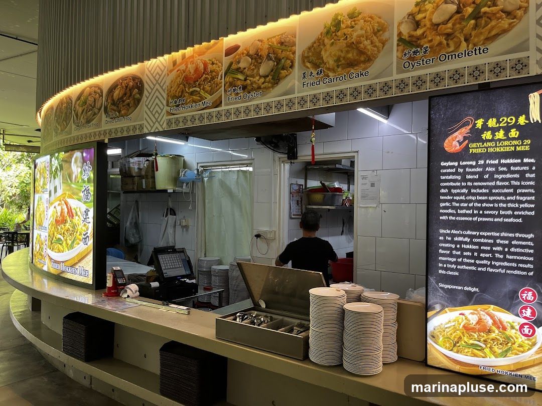 A food stall counter with various dishes displayed on a backlit menu and a descriptive sign. The stall appears to be serving Hokkien Mee and other noodle dishes. A staff member is visible in the background. Stacks of plates are neatly arranged on the counter.