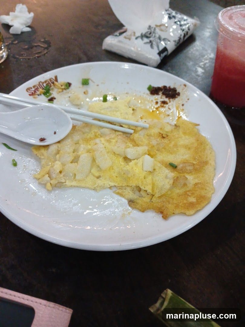 A white plate with leftover food, including an omelet and what appears to be rice cake or radish cake, is shown on a dark wooden table. Chopsticks and a spoon rest on the plate. A cup of red liquid, possibly a drink, and a packet of tissues are also visible. The plate has a logo or text that reads "Satay" on its rim.