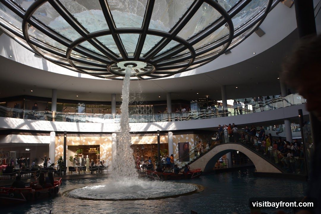 An indoor shopping mall features a large, multi-level fountain in the center, with water cascading from a circular structure above. Several boats are navigating the water. Shops, including Gucci and Tory Burch, are visible on the upper levels, with people browsing and walking around. The architecture is modern, with a glass ceiling and balconies.