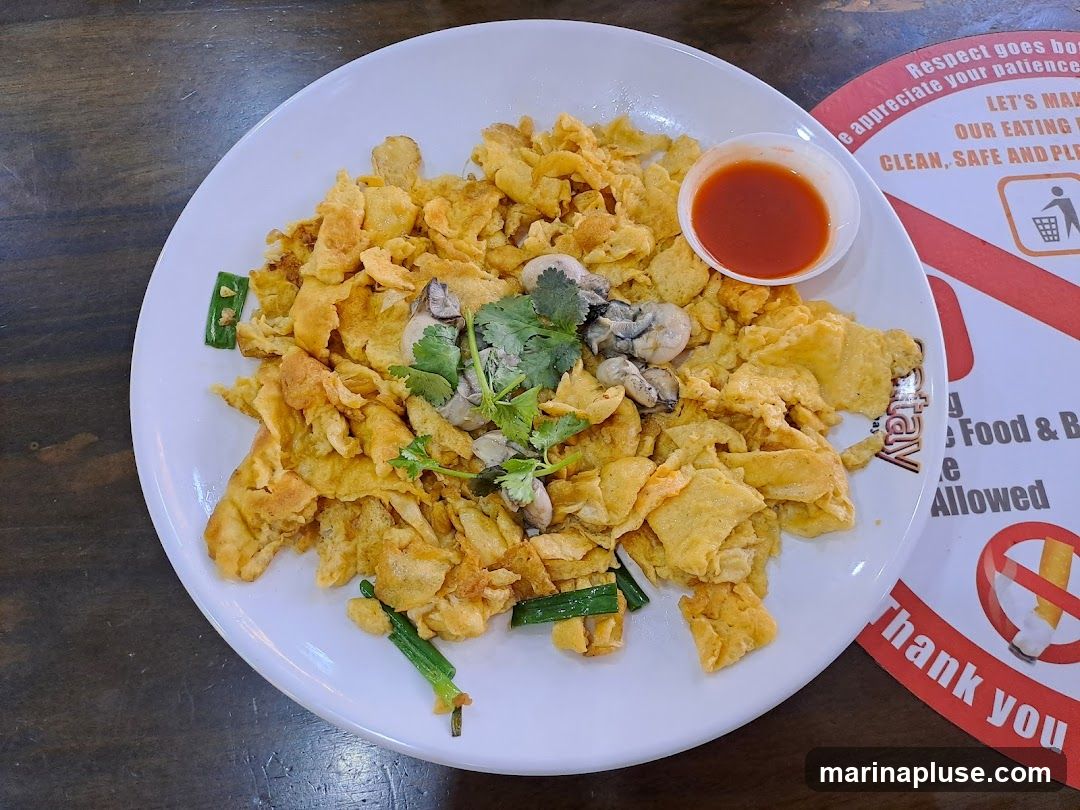 A plate of oyster omelet with a small dish of chili sauce and garnished with cilantro and green onions. A circular sign with text about cleanliness and a no-smoking symbol is visible on the right.