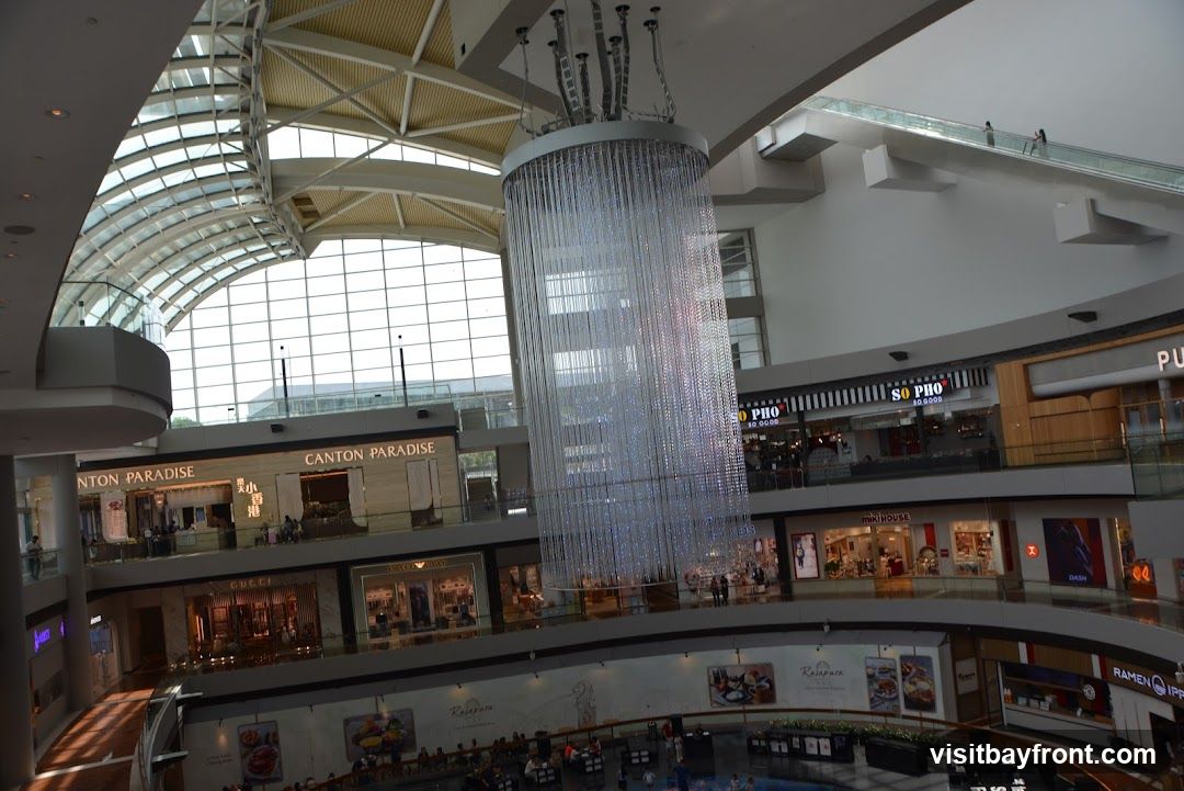 The image shows the interior of a multi-level shopping mall. A large, cylindrical chandelier made of numerous hanging strands of lights dominates the center of the frame. Various shops are visible on different levels, including 'CANTON PARADISE', 'SO PHO', 'GUCCI', 'ARMANI EXCHANGE', and 'RAMEN IPPUDO'. Escalators are also visible, with a few people on them. The mall has a modern architectural design with a curved glass ceiling.