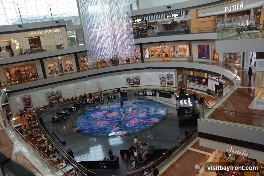 The image shows the interior of a large, modern shopping mall. A prominent feature is a large, cylindrical chandelier made of numerous lights, hanging in the center of the atrium. Below the chandelier, the ground floor features a circular area with a vibrant, illuminated display on the floor, where several people are walking. Surrounding this central area are various shops and restaurants on multiple levels, with balconies and walkways overlooking the atrium. Visible shop names include 'CANTON PARADISE', 'GUCCI', 'EMPORIO ARMANI', 'SO PHO SO GOOD', 'MIKI HOUSE', 'DASH', 'RAMEN IPPUDO', 'PUTIEN', and 'Venchi'. People are seen dining at tables on the lower levels and browsing in the shops.