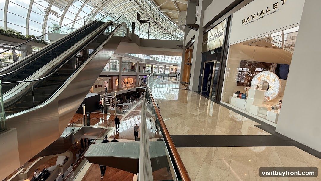 The image shows the interior of a multi-level shopping mall with escalators, shops, and walkways. The architecture features a large, curved glass roof allowing natural light to illuminate the space. On the right side of the image, a storefront with the name 'DEVIALET' is visible, displaying various products in its window. People are seen walking on different levels and using the escalators.