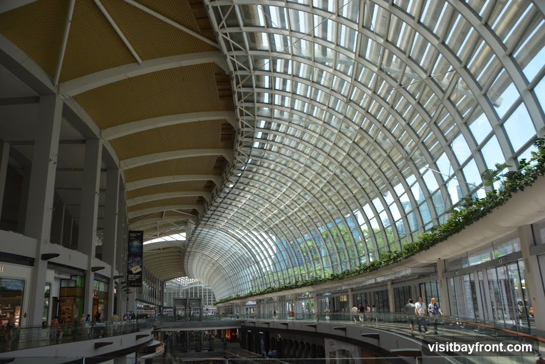An interior view of a large, modern shopping mall with a distinctive curved glass roof and a multi-level design. The architecture features prominent white columns and beams supporting a yellow-toned ceiling on one side, contrasting with the expansive glass structure on the other. Greenery is visible along the upper walkway, and people are seen browsing and walking on different levels. The overall impression is one of spaciousness and contemporary design, with natural light flooding the interior through the glass roof.