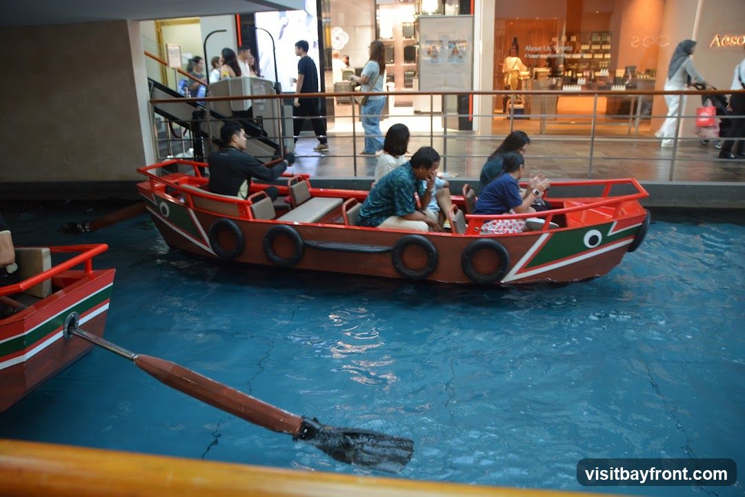 A group of people are enjoying a boat ride in a canal within a shopping mall. The boats are red and brown with green and white accents. Several individuals are seated in the boats, some rowing and others observing. In the background, shoppers can be seen walking past storefronts.