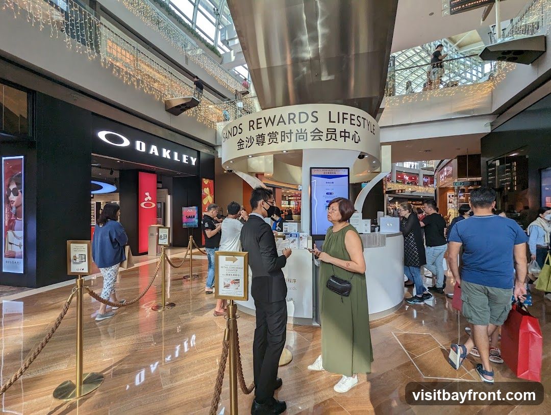 A view inside a modern shopping mall with a prominent Oakley store. People are seen browsing and queuing. A circular information desk with signage in English and Chinese is also visible. Decorative lights hang from the ceiling and railings.