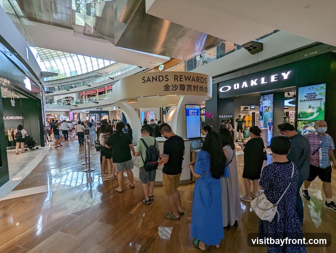 A wide shot of a busy shopping mall interior with various stores and shoppers. In the foreground, people are queuing near a 'Sands Rewards' kiosk. To the right, the Oakley store is visible with its prominent signage and displays of merchandise. Other shops and escalators can be seen in the background, with a large, modern ceiling structure overhead.