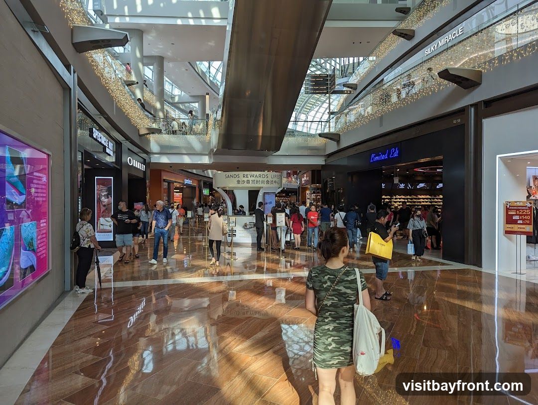 A bustling interior of a modern shopping mall with multiple levels. Stores like Oakley, Limited Edt, and Silky Miracle are visible. People are walking around, browsing shops, and interacting. Decorative lights are strung along the railings of the upper levels. A promotional sign for Sands Rewards is also present.