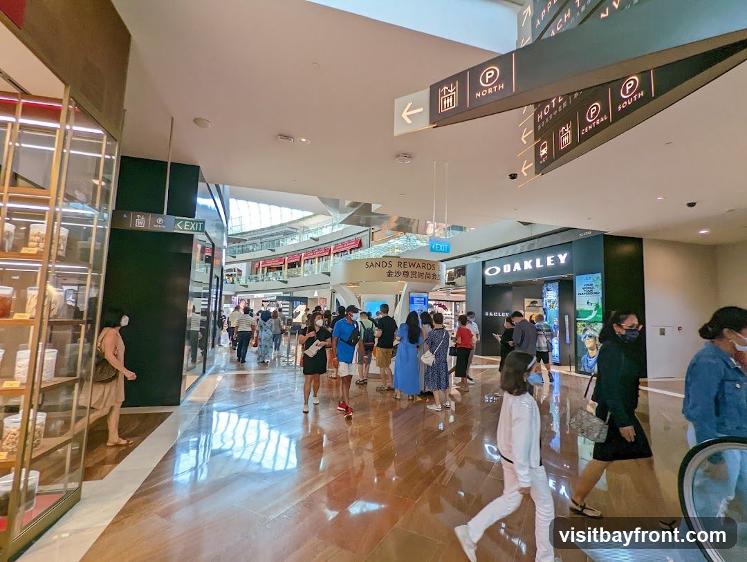 An interior view of a busy shopping mall with many shoppers. Signs indicate directions to North, South, Central, and Hotel. An Oakley store is visible with its prominent logo. To the left, a display shelf with jars of goods is seen.