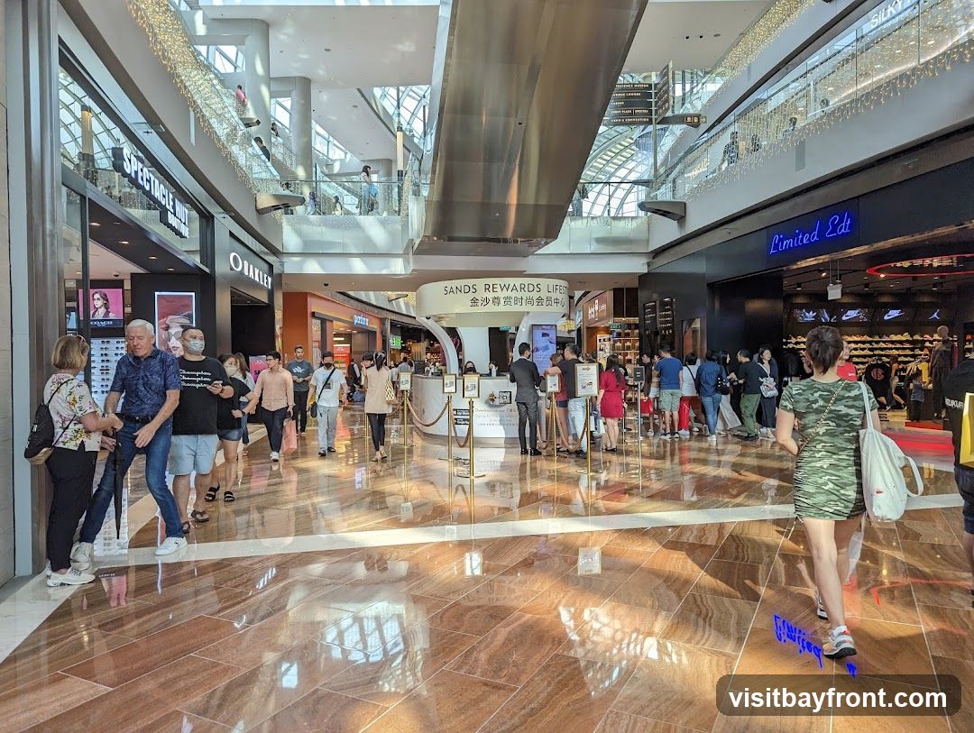 People are walking through a bright, spacious shopping mall with polished floors reflecting the overhead lights and architectural features. Several storefronts are visible, including Spectacle Hut, Oakley, and Limited Edt, which displays shoes. A central kiosk with signage for "Sands Rewards Lifestyle" is also present. The mall appears to be busy with shoppers browsing and moving between stores.