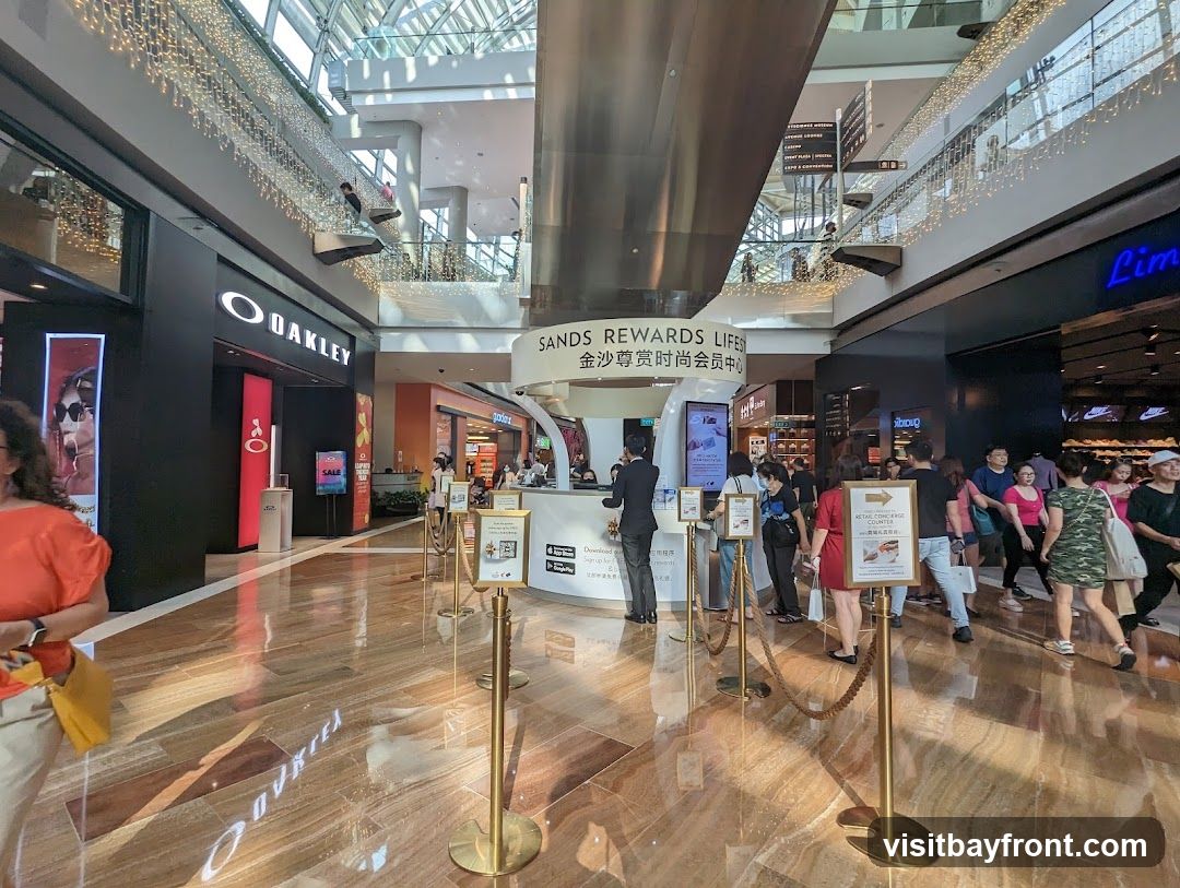 A wide shot of a busy shopping mall interior with various stores and shoppers. In the foreground, a polished marble floor reflects the overhead lights and store signs. A queue forms at a "Sands Rewards Lifescape" counter. To the left, an Oakley store is visible with its prominent logo. Further down the mall, other shops and escalators can be seen, adorned with decorative string lights.
