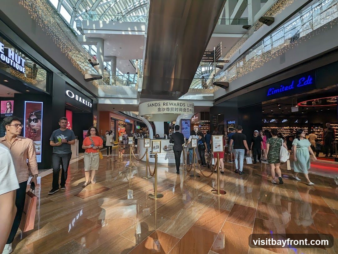 A wide shot of a busy shopping mall interior with multiple stores visible. People are walking around the polished floor. Stores like Oakley and Limited Edt are identifiable. Decorative lights are strung along the upper levels of the mall. A "Sands Rewards Life" counter is also visible.