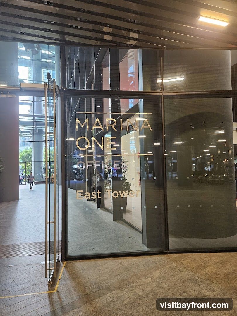 A glass entrance with gold lettering that reads "MARINA ONE OFFICES East Tower". The doors are open, revealing a walkway and people inside.