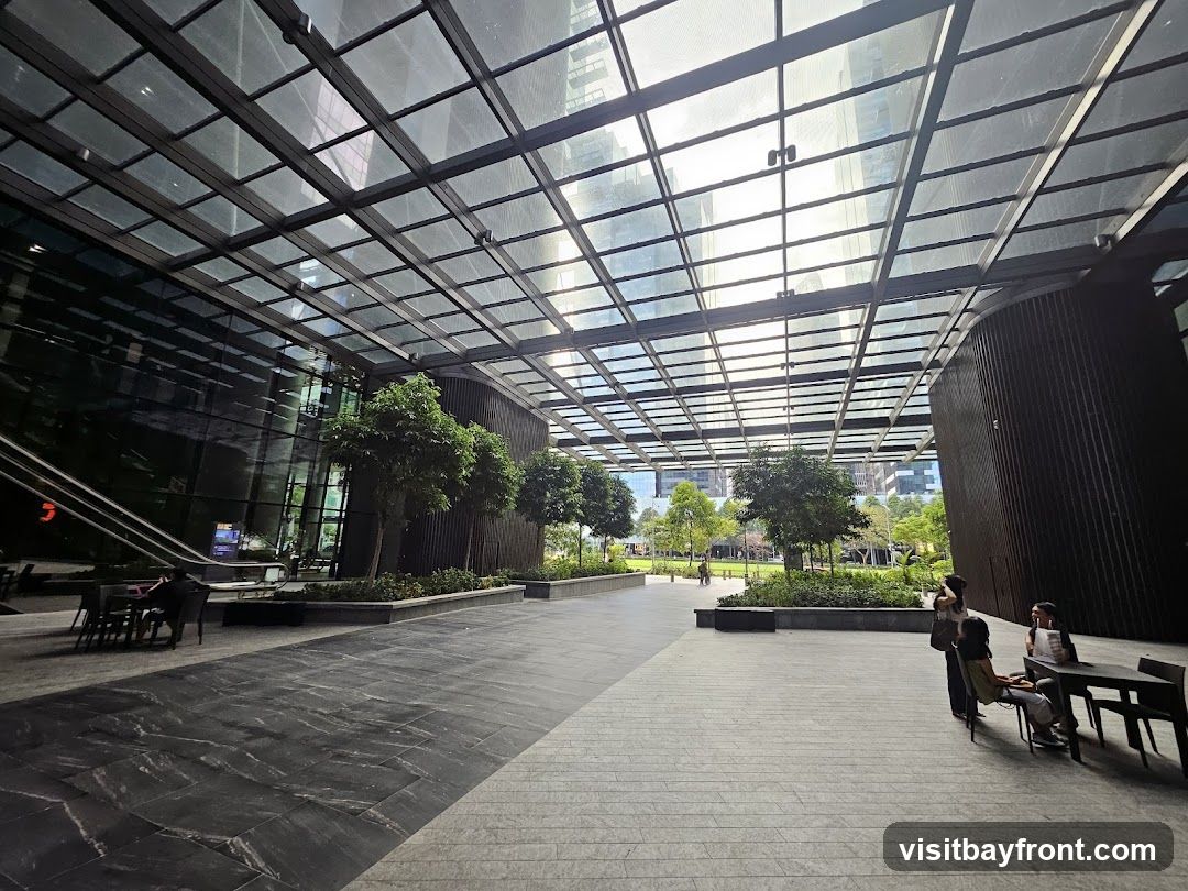 An expansive outdoor plaza features a sleek, modern design with a large glass canopy overhead, allowing natural light to filter through. The area is furnished with tables and chairs, suggesting a space for relaxation or dining. Lush greenery, including trees and manicured garden beds, is interspersed throughout the plaza, creating a pleasant, urban oasis. People are seen casually interacting and seated at tables, enjoying the open space. The architecture of the surrounding buildings is visible through the glass canopy, reflecting the sky and urban landscape.