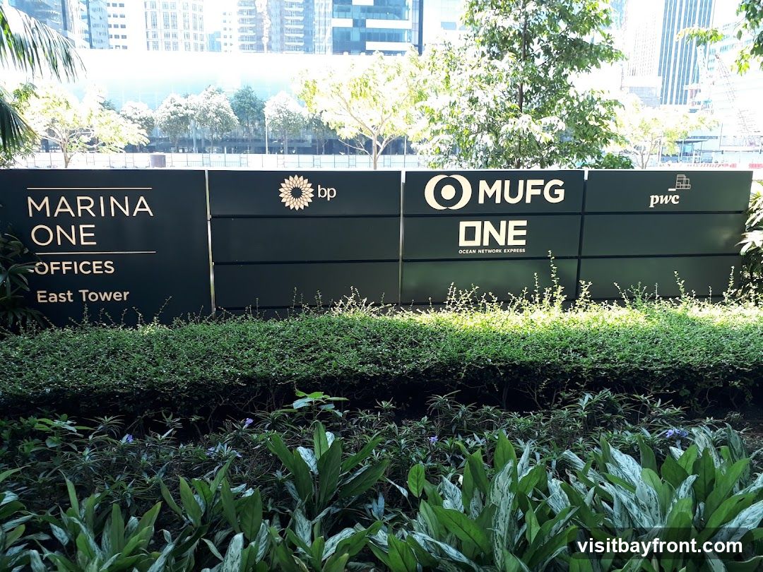 A dark green sign with white lettering displays the names of various businesses and offices. The sign reads "MARINA ONE OFFICES East Tower" on the left. To the right, it shows the logos and names "bp", "MUFG ONE OCEAN NETWORK EXPRESS", and "pwc". The sign is set against a backdrop of lush green bushes and trees, with modern buildings visible in the distance under a bright sky.