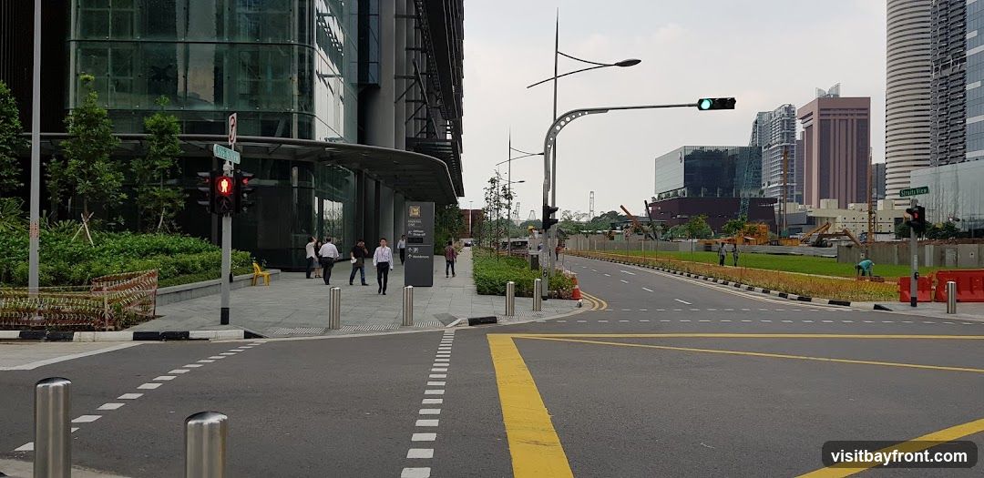 A wide, paved street in a modern city is depicted. On the left, a contemporary building with a glass facade and dark metal accents stands tall. A pedestrian walkway runs alongside the building, with several people walking. Traffic lights are visible, showing red for pedestrians and green for vehicles. Lush green landscaping with trees and bushes borders the walkway. On the right side of the street, more modern buildings rise into the sky, some appearing to be under construction with cranes and scaffolding. The road has clear lane markings, including a prominent yellow line. The sky is overcast.