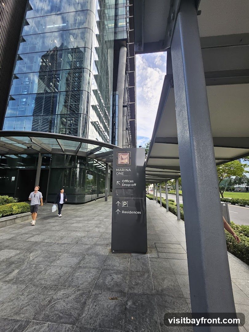 A tall, modern glass building with reflections of other buildings and the sky. A covered walkway runs along the side of the building, with a dark gray signpost indicating directions to