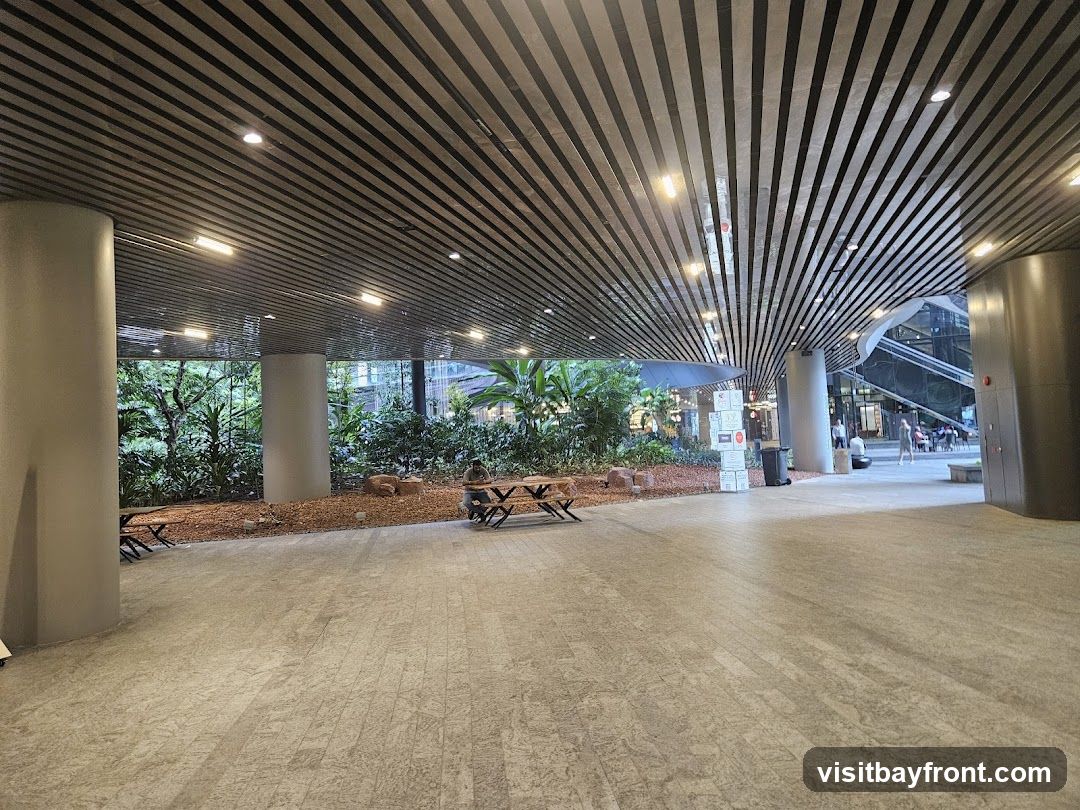 An outdoor seating area with wooden tables and benches is visible, surrounded by lush greenery. The area features a modern ceiling with parallel black and white slats and recessed lighting. In the background, people are seen walking and sitting at tables, suggesting a public space or commercial area.