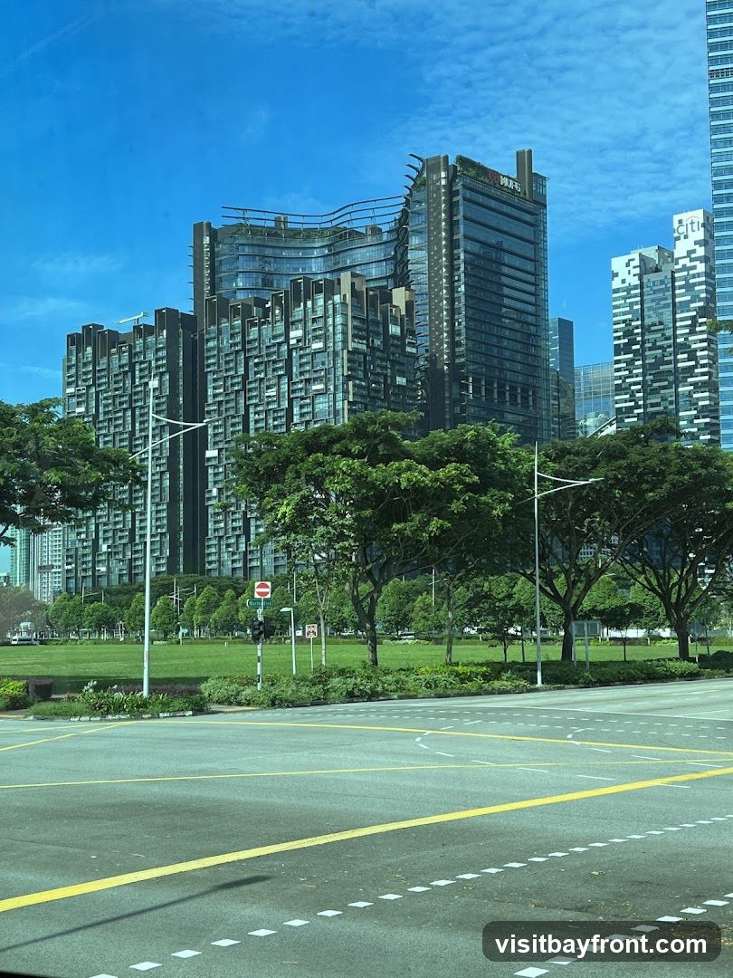 A view of a modern cityscape featuring several tall buildings with unique architectural designs. In the foreground, there is a road intersection with clear lane markings. Behind the road, a large, well-maintained green lawn is visible, dotted with trees. The sky is bright blue with scattered clouds. One of the buildings has visible signage that appears to read 'CITI'.