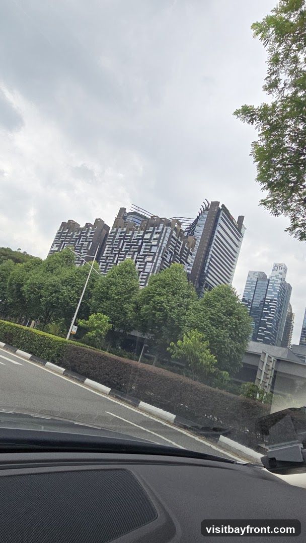A view of contemporary high-rise buildings with distinctive architectural designs, set against a backdrop of lush green trees and a cloudy sky. The buildings feature a mix of glass facades and patterned exteriors. A road and a manicured hedge are visible in the foreground, suggesting an urban or suburban setting.