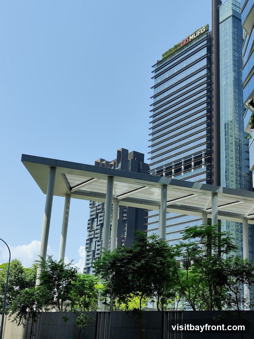 A view of modern, glass-facade office buildings under a clear blue sky. The building in the center has a green roof and the MUFG logo is visible at the top. In the foreground, there is a covered walkway supported by large pillars, with lush green trees and foliage below.