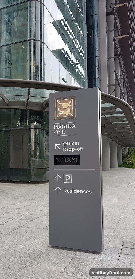 A tall, gray directional sign stands on a paved walkway in front of a modern glass building. The sign displays the "Marina One" logo at the top, followed by directional arrows and text indicating "Offices Drop-off," "TAXI," and "Residences." An arrow points left for offices and drop-off, a black illuminated box with an arrow points left for taxi, and two upward arrows indicate parking and residences. The building behind the sign has a reflective glass facade with horizontal architectural elements.