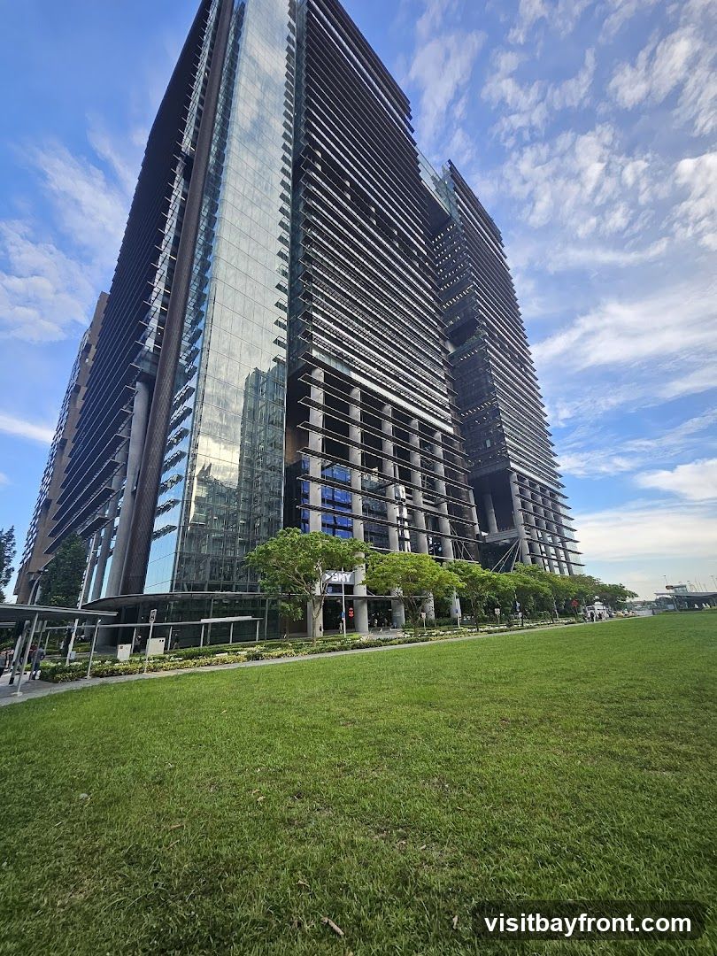 A low-angle shot of a tall, modern skyscraper with a facade of glass and horizontal louvers. The building is set against a bright blue sky with scattered white clouds. In the foreground, a lush green lawn stretches out, with a paved walkway and some trees lining the base of the building. The overall scene conveys a sense of urban development and open space.