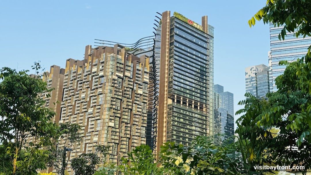 A view of several modern skyscrapers with unique architectural designs, partially obscured by lush green trees and foliage in the foreground. One of the buildings features a prominent red logo with the text "MUFG" at its top. The sky is clear and blue.