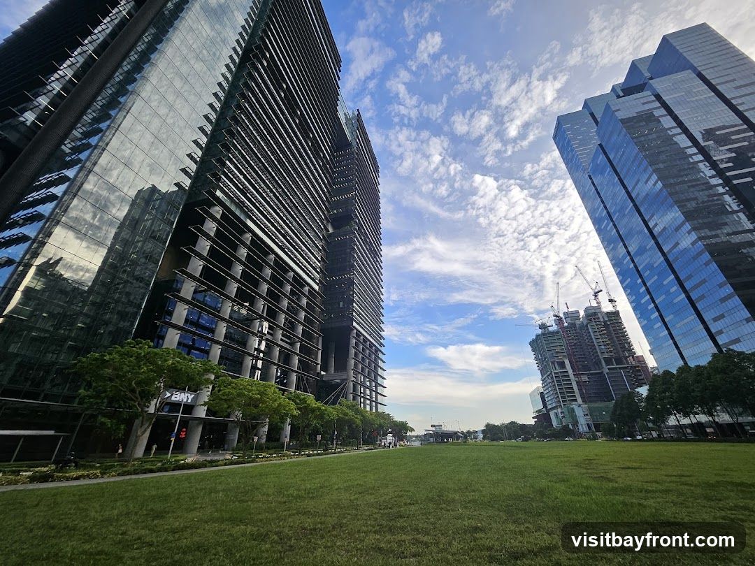 A wide-angle shot captures a modern cityscape dominated by towering glass skyscrapers under a bright, cloudy sky. In the foreground, a lush green lawn stretches out, bordered by trees and a pathway. The buildings feature sleek, reflective facades, with one prominently displaying the 'BNY' logo. In the background, construction cranes indicate ongoing development. The scene conveys a sense of urban development and open green spaces within a city.