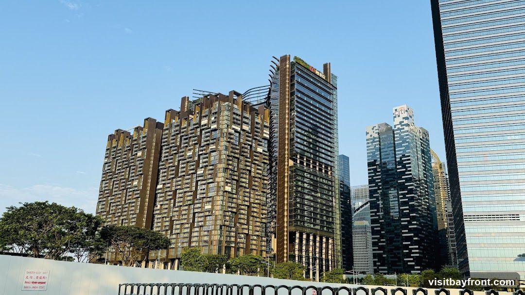 A wide-angle view of a modern cityscape dominated by several tall skyscrapers. The buildings feature diverse architectural styles, with one prominent building showcasing a unique, tiered design with a metallic facade. Another skyscraper has a sleek, glass exterior, reflecting the clear blue sky. In the foreground, a white barrier with some signage and a row of black metal fencing is visible. Trees and greenery are interspersed between the buildings and along the foreground. The overall scene depicts a bustling urban environment under a bright sky.