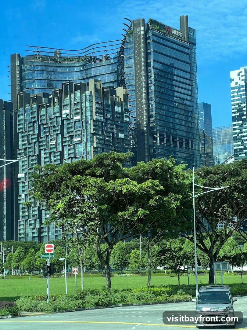 A view of contemporary skyscrapers with unique architectural designs, set against a bright blue sky. In the foreground, lush green trees and manicured lawns create a natural contrast to the urban environment. A silver van is visible on the road in the lower right corner. A street sign indicates 'Marina Blvd'. The buildings feature glass facades and intricate structural elements, with one prominent building displaying the MUFG logo at its apex.