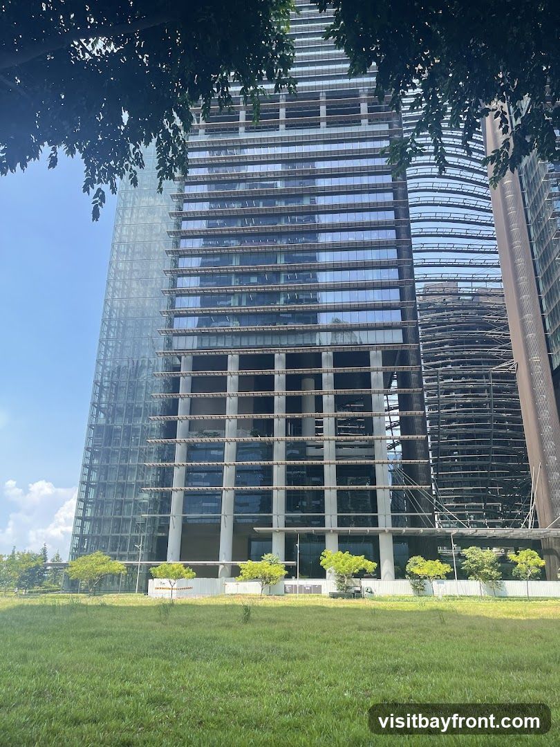 A tall, modern skyscraper with a glass facade stands under a clear blue sky. The building features horizontal bands of windows and structural elements. In front of the building, there is a strip of green grass with several small trees. The lower portion of the building appears to be an entrance area, but no specific business is identifiable.